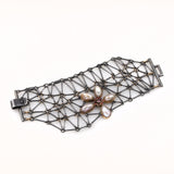 Oxidized Sterling silver chain mail bracelet made of rods and rings. The round connecting rings are 18KY gold. There is a large flower motif on the edge of the bracelet. The border of the bracelet is irregular and the strap lock is visible. The center of the flower is made of bezel set rhodolite garnets and orange sapphires.