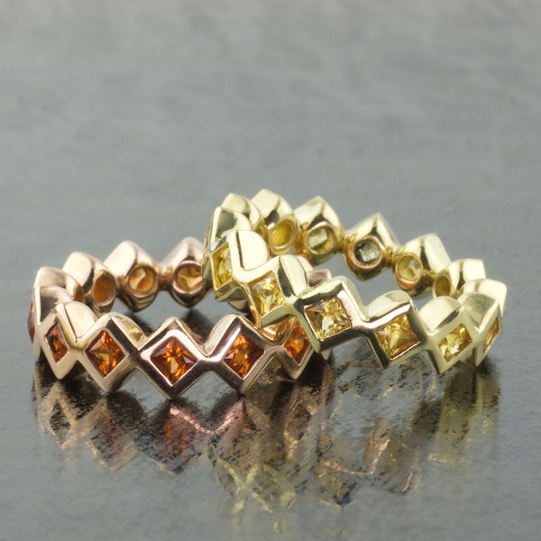 Close up of two zig zag bands Eternal Bands; one in 18KY gold set with Princess-cut yellow Sapphires and the other in 14KR gold with orange Princess-cut Sapphires